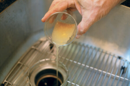 person pouring cloudy liquid from a plastic cup slowly in to a kitchen sink drain